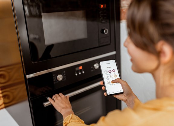 Smart Home Frau mit Smartphone in Küche vor Backofen mit Home Connect